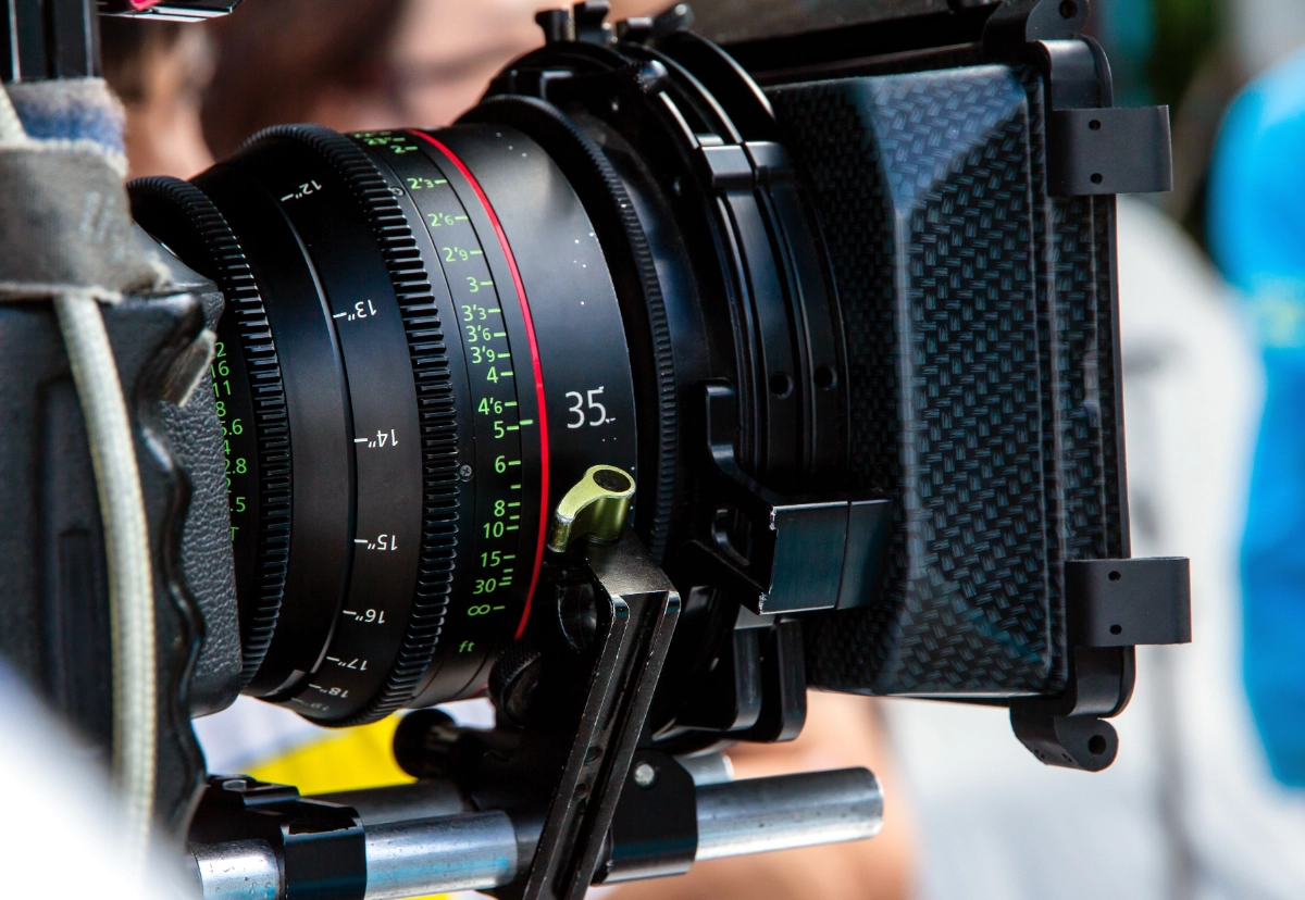Na zdjęciu znajduje się profesjonalna kamera filmowa wyposażona w wysokiej klasy obiektyw oraz matte box. Widoczne są precyzyjne oznaczenia ostrości i ogniskowej, charakterystyczne dla sprzętu wykorzystywanego podczas produkcji filmów, reklam i animacji. Fotografia przedstawia zaplecze techniczne profesjonalnej realizacji wideo.