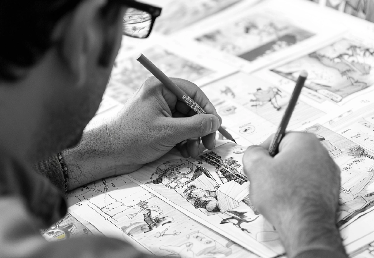 Na zdjęciu widać osobę szkicującą storyboard do animacji. Na stole znajdują się kartki z odręcznymi rysunkami przedstawiającymi kolejne kadry filmu, a twórca dopracowuje ilustracje za pomocą ołówków.